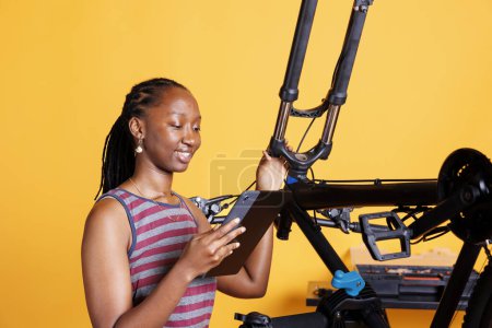 Photo for Young black lady inspecting damaged bicycle and exploring repair options on her smart tablet. Image of african american woman checking broken bike components while holding a digital device. - Royalty Free Image