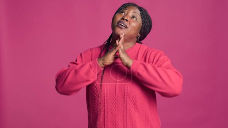 Jovencita de etnia afroamericana de pie y rezando con fidelidad sobre fondo rosa. Mujer negra en estilo elegante mirando hacia arriba con las manos arriba mostrando devoción.