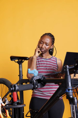 Femme noire dédiée répare expertement vélo cassé en utilisant des outils spécialisés et la technologie sans fil pour la recherche et les instructions. Jeune femme vibrante surfant sur Internet pour l'entretien des engins de vélo.
