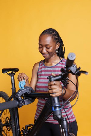 Foto de Mujer ciclista negra inspeccionando su marco roto de bicicleta mientras se coloca en el soporte de reparación contra un telón de fondo aislado. Mujer joven que se prepara para utilizar herramientas especializadas para ajustes y reparaciones. - Imagen libre de derechos