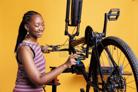 Téléchargez les photos : Une jeune cycliste active veille à ce que le vélo soit bien préparé pour les aventures en plein air en examinant attentivement les pédales et l'équipement. Femme africaine américaine sportive saisissant le levier de pied de vélo pour l'entretien. - en image libre de droit