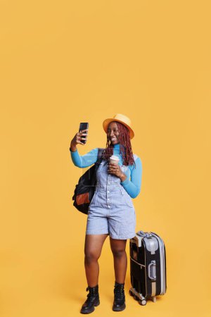 Photo for Traveller taking photos on vacation trip, making silly faces on smartphone and drinking coffee. Female person having fun with pictures in studio, carrying backpack and trolley bags. - Royalty Free Image