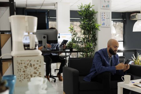 Foto de Trabajador de negocios árabe charlando con su compañero de trabajo usando un teléfono inteligente mientras trabaja en un espacio abierto moderno. Marketing profesional sentado en el sofá y mensajes de texto en las redes sociales en el teléfono móvil en la oficina de inicio - Imagen libre de derechos