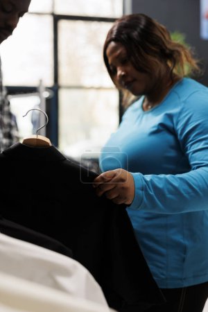 Téléchargez les photos : Femme élégante vérifiant matériau chemise, en regardant les cintres avec des vêtements à la mode dans la boutique moderne. Afro-américain client shopping pour l'usure formelle, l'achat d'articles élégants - en image libre de droit
