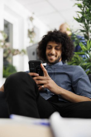 Photo for Smiling businessman relaxing in office, using smartphone to send message. Young arab executive manager texting in social media on mobile phone while having break in coworking space - Royalty Free Image