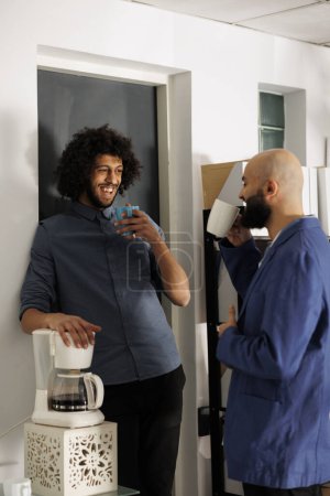 Two smiling arab businessmen having coffee break and talking in business office. Company employees standing with tea mugs while laughing and chatting in coworking space workplace