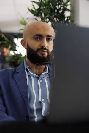 Téléchargez les photos : Homme d'affaires arabe assis devant un ordinateur portable au bureau, se préparant à un appel vidéo avec des clients. Directeur exécutif vérifiant le rapport d'analyse financière avant sa présentation dans un espace de coworking - en image libre de droit