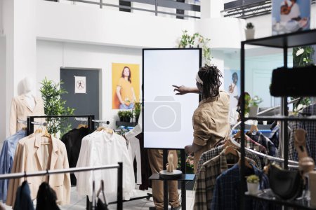Téléchargez les photos : Homme afro-américain tout en utilisant un panneau d'affichage interactif intelligent dans un magasin de vêtements. Client tapotant sur l'écran blanc vierge maquette pour chaussures annonces numériques et des informations dans le centre commercial boutique - en image libre de droit