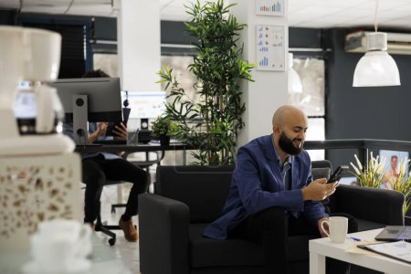 Téléchargez les photos : Entrepreneur souriant répondant à un message texte sur smartphone dans le bureau de démarrage. Jeune homme d'affaires arabe utilisant les médias sociaux sur un téléphone mobile tout en ayant pause au travail - en image libre de droit