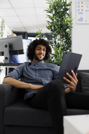 Téléchargez les photos : Arabe employé d'affaires télétravail, bavarder en réunion en ligne à l'aide d'une tablette numérique. Sourire détendu start-up entrepreneur visioconférence tout en étant assis sur le canapé dans le bureau - en image libre de droit