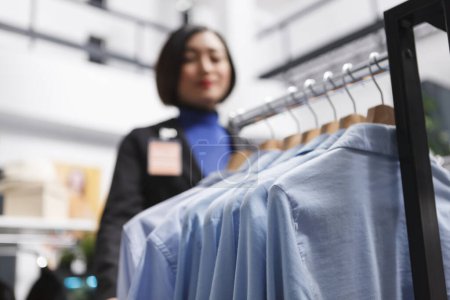 Foto de Camisas formales colgando en estante de exhibición, mientras que la mujer asiática asistente de trabajo en la tienda de ropa enfoque selectivo. Ropa de hombre prenda en perchas en el centro comercial boutique primer plano - Imagen libre de derechos