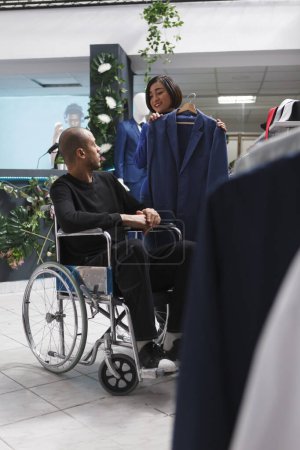 Photo for Clothing store asian woman employee providing support to customer with physical impairment in selecting jacket. Mall worker consulting arab man in wheelchair in choosing formal outfit - Royalty Free Image