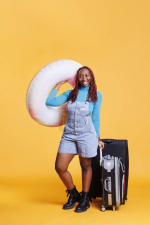 African american girl leaving on holiday with inflatable ring and trolley bags, feeling excited about vacation. Young woman enjoying weekend activities abroad, carrying luggage.