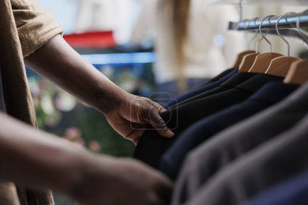 Téléchargez les photos : Homme afro-américain naviguant vestes sur rack tout en choisissant les vêtements masculins formels dans le magasin de vêtements. Acheteur mains à la recherche de blazer élégant accroché dans le centre commercial boutique de mode - en image libre de droit