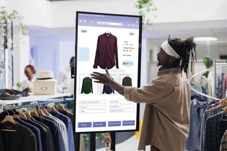 Afrikanisch-amerikanischer Kunde wählt Kleidung auf Touchscreen-Board im Bekleidungsgeschäft aus und kauft Artikel an der Selbstbedienungskasse. E-Commerce-Selbstbestellkonzept in der Modeboutique.