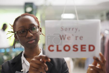 Téléchargez les photos : Magasin de vêtements femme afro-américaine manager pendre désolé que nous sommes fermés signe sur la fenêtre. Assistant de service de centre commercial de vêtements tenant signe remarquant des clients au sujet de la fin des heures d'ouverture - en image libre de droit