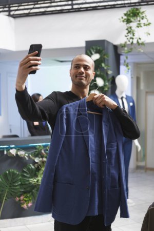 Foto de Hombre árabe alegre tomando selfie y mostrando equipo usando la cámara frontal del teléfono inteligente durante las compras en la boutique. Blogger sostiene chaqueta y transmisión en vivo en el teléfono móvil en la tienda de ropa - Imagen libre de derechos
