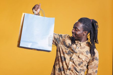Photo for African american shopper man holding shopping bags in hands after buying stylish clothes. Shopaholic young adult smiling at camera after purchase fashion items in studio over yellow background - Royalty Free Image