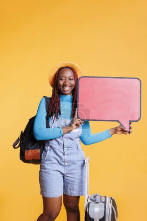 Photo for Happy tourist with speech bubble icon creating ad, travelling on holiday trip with bags. Young female model showing copyspace empty cardboard with advertisement in studio, isolated board. - Royalty Free Image