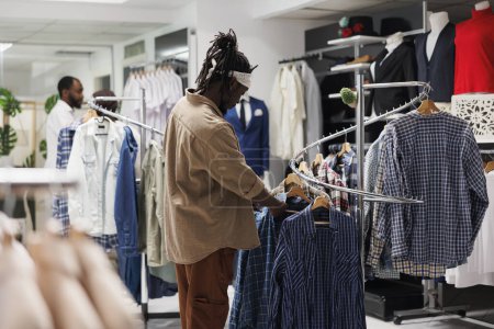 Foto de Cliente afroamericano mirando camisas colgantes en estante en boutique de moda. Almacene ropa de control de clientes en perchas mientras elige ropa casual de moda en el centro comercial - Imagen libre de derechos