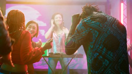 Foto de Diverso grupo de amigos de fiesta en la pista de baile, DJ mujer mezclando sonidos en el escenario en la discoteca. Gente alegre bailando música funky en el club nocturno, divirtiéndose con luces brillantes. - Imagen libre de derechos