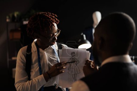 Foto de Diseñador de moda experimentado mostrando dibujo bosquejo couturier de vestido de novia que ella quiere que él personalice. modistas afroamericanos trabajando en elegante traje sartorial a medida - Imagen libre de derechos