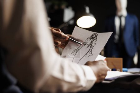 Photo for Meticulous expert dressmaker designing comissioned wedding dress for customer in luxury atelier shop. Skilled tailor looking over sketch drawing, planning upcoming adjustments - Royalty Free Image
