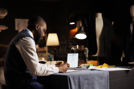 Téléchargez les photos : Un gentleman afro-américain esquisse à la main la prochaine collection de restaurants sur mesure dans un atelier de couture. Créateur de mode expert créant pièce de vêtement créative sur mesure - en image libre de droit