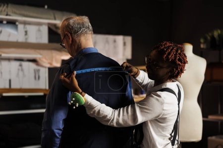 Photo for Expert seamstress sizing up stylish client proportions for upcoming luxurious bespoke costume. Elderly customer getting measurements taken by meticulous suitmaker in tailoring studio - Royalty Free Image