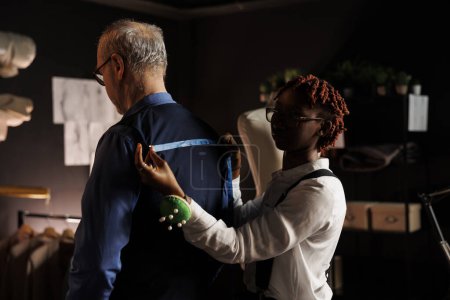 Photo for Senior gentleman getting back measurements taken by precise seamstress in tailoring studio. Professional couturier sizing up client proportions for custom made vest comission - Royalty Free Image