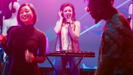Cool woman mixing sounds at DJ station, creating funky atmosphere at nightclub on stage. Cheerful modern girl putting music on audio panel, partying and clubbing with people on dance floor.