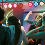 Crowd of people jumping on music, feeling happy on dance floor at nightclub. Young funky friends dancing on electronic music with DJ on stage, partying together with spotlights.