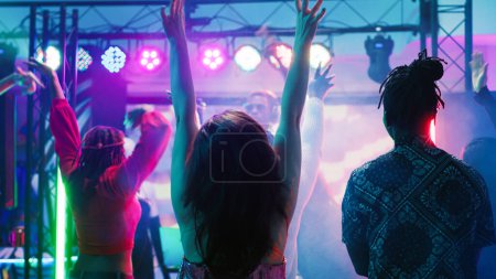 Photo for Diverse people partying at nightclub, enjoying live music performance from DJ station at discotheque stage. Group of friends dancing together on dance floor with spotlights. Tripod shot. - Royalty Free Image
