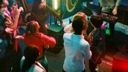 Téléchargez les photos : Des hommes et des femmes joyeux faisant la fête ensemble sur la piste de danse, écoutant de la musique électronique de DJ mixer station sur scène. Diverses personnes dansent et sautent dans la discothèque. Coup de main. - en image libre de droit