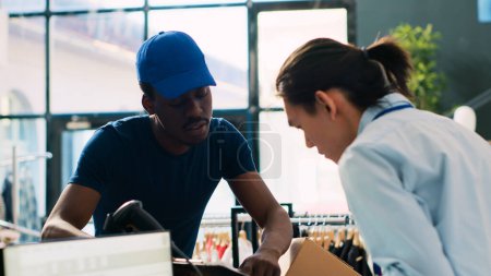 Photo for Asian worker signing packages delivery report, discussing clothes distribution with african american courier in modern boutique. Store manager preparing boxes with customers orders in shopping centre - Royalty Free Image
