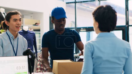 Photo for Courier in delivery uniform in modern boutique, discussing packages shipping details with store manager. African american deliveryman showing distribution report on tablet computer. Fashion concept - Royalty Free Image