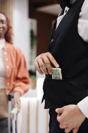 Photo for Bellboy accepting cash from people, putting money in pocket and carrying suitcases for hotel guests. Male steward working as concierge taking tip from tourists, luxury service. Close up. - Royalty Free Image