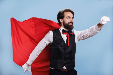 Strong superhero character with red cape posing against blue background, feeling confident and determined in front of camera. Young man impersonating hero with mantle, shows force.