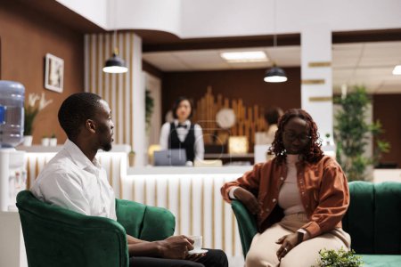 Téléchargez les photos : Les clients afro-américains dans le salon parlent et profitent d'un service de luxe à l'hôtel, attendant de remplir le formulaire d'inscription pour la réservation de chambre. Touristes assis sur le canapé à la réception. - en image libre de droit