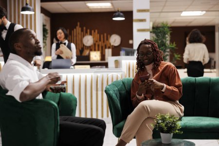 Photo for African american woman using smartphone sitting in lounge area at hotel, preparing for room accommodation at luxury resort. Couple waiting to do check in process at reception counter. - Royalty Free Image