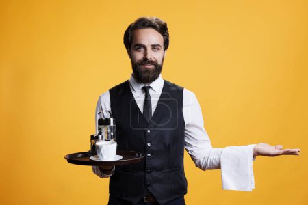 Photo for Gracious waiter creating marketing ad in studio, posing with tray in hand and presenting something to left or right sides. Young man working as luxurious butler serving people, elegant personnel. - Royalty Free Image