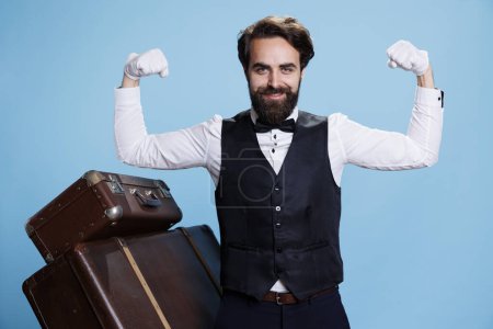 Photo for Professional porter flexing muscles on camera, showing off power and strength underneath his formal clothes. Hotel concierge employee feeling powerful in studio, hospitality industry. - Royalty Free Image