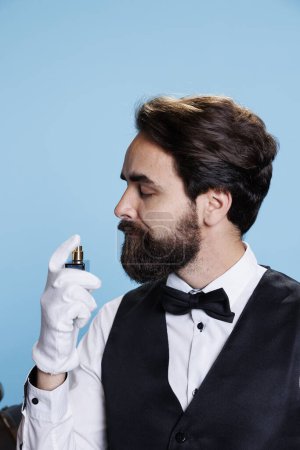 Photo for Hotel porter using cologne bottle to smell nice, applying perfume on camera and feeling confident while he wears professional formal suit with tie. Employee spraying fresh aromatic scent. Close up. - Royalty Free Image