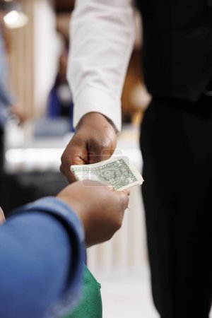 Photo for Close up of male African American waiter receiving tip from generous hotel guest. Tourist giving money cash to restaurant worker, making order, paying bill. Tipping in hospitality industry - Royalty Free Image