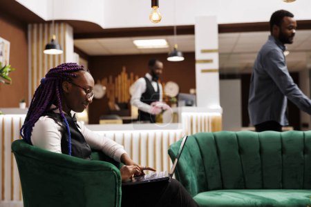 Jeune femme afro-américaine gestionnaire d'hôtel est assis dans le hall avec ordinateur portable en utilisant, examiner les check-ins et check-outs, garder une trace de l'inventaire et commander des fournitures