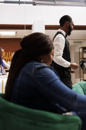Téléchargez les photos : Le personnel de l'hôtel afro-américain et invité dans le hall. Femme voyageuse en attente d'enregistrement à la réception, sympathique groom souriant aidant les clients avec les bagages. Hôtellerie et tourisme Industrie - en image libre de droit