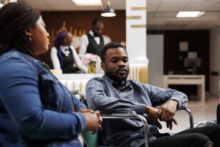 Young African American man in wheelchair in hotel lobby with able-bodied girlfriend, waiting for accessible room, handicapped guy traveling with mobility impairment. Accommodations for disabled