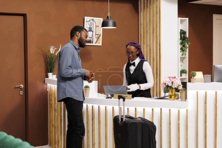 Friendly smiling African American female receptionist talking with hotel guest at front desk, checking his passport. Black guy traveler standing with suitcase at reception during check-in procedure