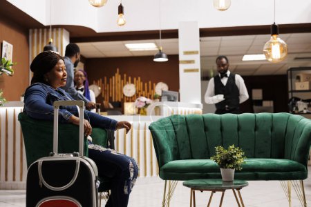Téléchargez les photos : Une voyageuse afro-américaine fatiguée assise avec des bagages dans le hall de l'hôtel attend le processus d'enregistrement, se sentant décalée par rapport au jet. Touriste féminine est arrivée à la station, attendant la chambre. Les gens et les voyages - en image libre de droit