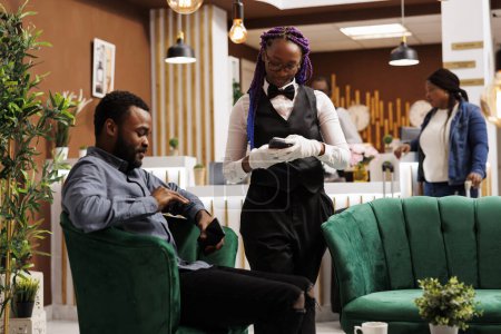 Téléchargez les photos : Homme afro-américain touriste assis dans le hall de l'hôtel payer pour le café avec smartphone, effectuer un paiement sans contact. Serveuse sympathique tenant pos terminal desservant les clients au salon du complexe de luxe - en image libre de droit